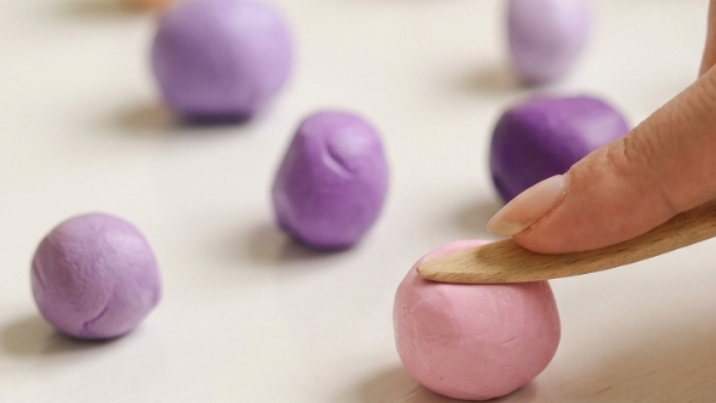 A close-up of a hand using a wooden clay tool to shape a pink dough ball, with several small colorful clay balls in purple and peach scattered on a light surface.