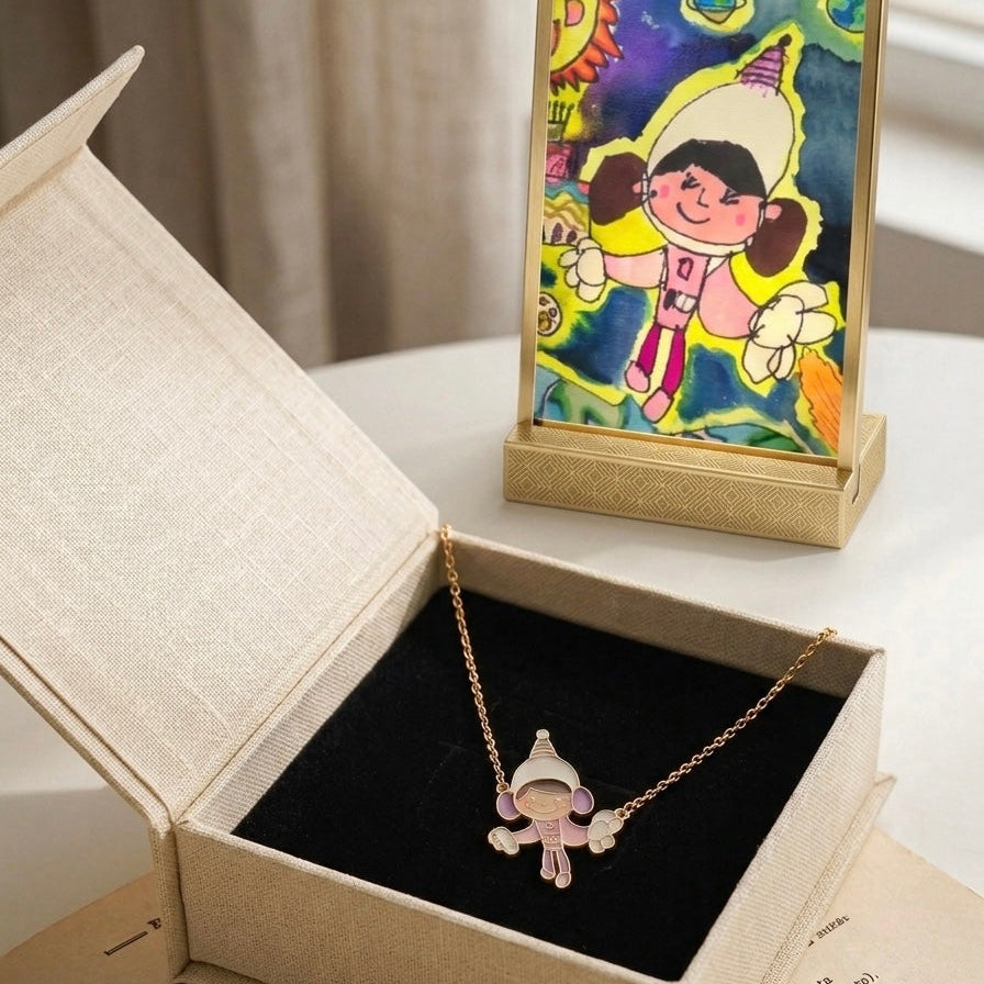 A gold accessories with a small cartoon character pendant displayed in an open beige jewelry box, with a colorful framed art print standing behind it on a light surface.