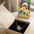 A gold accessories with a small cartoon character pendant displayed in an open beige jewelry box, with a colorful framed art print standing behind it on a light surface.