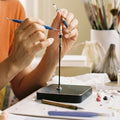 A close-up lifestyle shot of a person's hands using two blue-handled sculpting tools on a small jewelry stand over a worktable filled with craft supplies and pastel clay pieces.