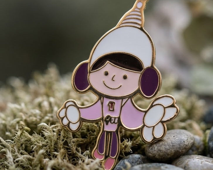 A small enamel pin shaped like a fairy or angel child, standing upright on moss and stones. The pin has a white hat, pink clothing, purple ear muffs and boots, and gold metal outlines, photographed outdoors with blurred rocks and moss in the background.