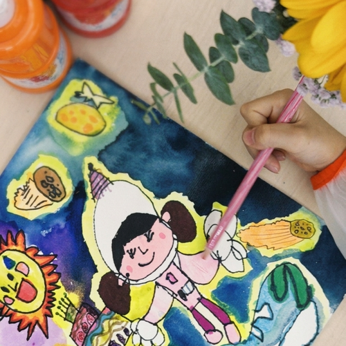 A close-up of a colorful children's painting on a canvas, showing cartoon astronauts and planets in bright blue, yellow, green, and orange tones. A hand holds a pink paintbrush over the artwork, with paint bottles and flowers visible around the painting setup.
